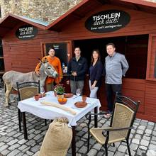 Eine Holzhütte, auf einem Schild im Giebel &#34;Tischlein deck Dich&#34;. Davor stehen ein Esel und 3 Männer und eine Frau und schauen in die Kamera. Davor ein gedeckter Tisch mit 2 Stühlen und einem Knüppel und Sack. ©Landkreis Saalekreis